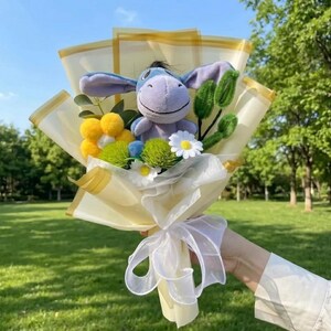 K&ouml;nnte beinhalten: Ein Blumenstrau&szlig; mit einem hellvioletten Pl&uuml;sch-Esel mit langen Ohren, umgeben von gelben und wei&szlig;en Stoffblumen, gr&uuml;nen Pompons und gr&uuml;nen Pfeifenreinigern. Das Arrangement ist in durchscheinendes cremefarbenes Papier mit wei&szlig;em Band gewickelt.