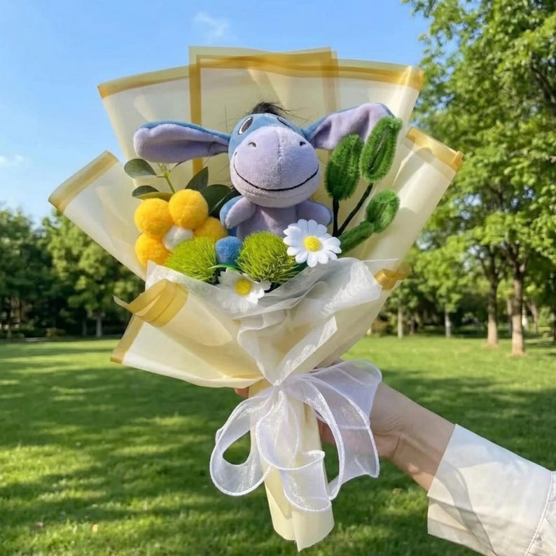 K&ouml;nnte beinhalten: Ein Blumenstrau&szlig; mit einem hellvioletten Pl&uuml;sch-Esel mit langen Ohren, umgeben von gelben und wei&szlig;en Stoffblumen, gr&uuml;nen Pompons und gr&uuml;nen Pfeifenreinigern. Das Arrangement ist in durchscheinendes cremefarbenes Papier mit wei&szlig;em Band gewickelt.