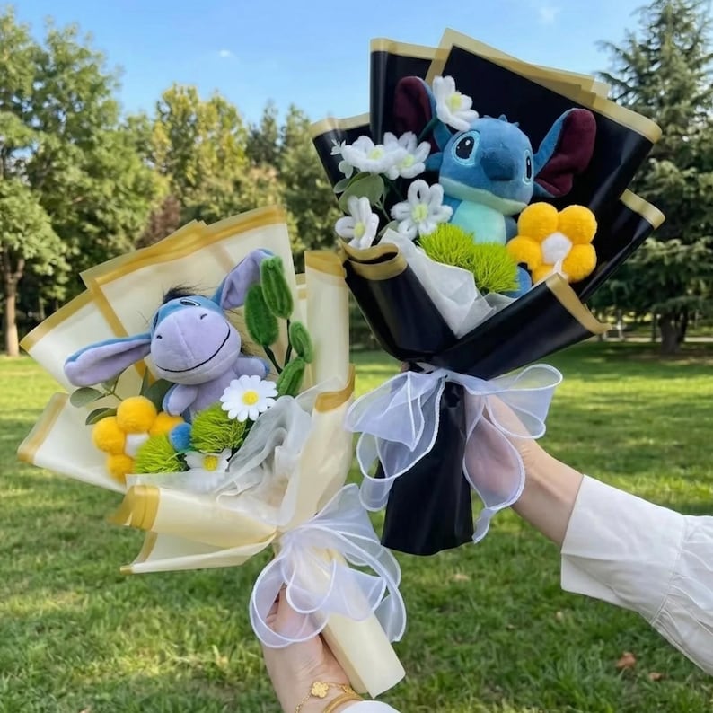 K&ouml;nnte beinhalten: Zwei Blumenstr&auml;u&szlig;e mit Pl&uuml;schfiguren. Ein Strau&szlig; enth&auml;lt einen hellvioletten Esel mit gelben und wei&szlig;en Blumen in cremefarbenem Papier. Der andere enth&auml;lt ein blaues Alien mit wei&szlig;en und gelben Blumen in schwarzem Papier.