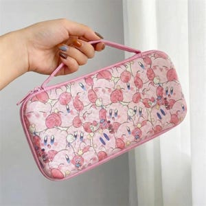 Può includere: Custodia da trasporto rosa con cerniera con un motivo ripetuto di personaggi dei cartoni animati. La custodia ha una maniglia e una cerniera rosa. I personaggi sono rosa con accenti bianchi e blu. Per una console di gioco.