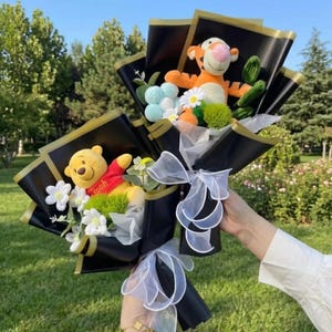 K&ouml;nnte beinhalten: Zwei Blumenstr&auml;u&szlig;e in schwarzem Papier mit Goldrand, jeweils mit einer Pl&uuml;schfigur. Ein Strau&szlig; enth&auml;lt einen gelben B&auml;ren in rotem Hemd, der andere einen orangefarbenen gestreiften Tiger. Beide enthalten wei&szlig;e Blumen und gr&uuml;ne Akzente.