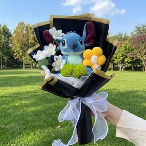 K&ouml;nnte beinhalten: Ein Blumenstrau&szlig; mit einer blauen Pl&uuml;schfigur mit gro&szlig;en Ohren, umgeben von wei&szlig;en und gelben Stoffblumen sowie gr&uuml;nen, strukturierten Akzenten. Das Arrangement ist in schwarzes Papier mit Goldrand gewickelt und mit einem wei&szlig;en Schleifenband versehen.