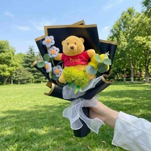 K&ouml;nnte beinhalten: Ein Winnie Puuh Pl&uuml;schtier in einem roten T-Shirt mit dem aufgestickten Namen Pooh, arrangiert als Blumenstrau&szlig; mit wei&szlig;en Filzblumen, gr&uuml;nen Pompons und Eukalyptusbl&auml;ttern, eingewickelt in schwarzes Papier mit goldenem Rand und wei&szlig;em Band.