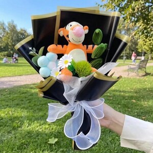K&ouml;nnte beinhalten: Ein Blumenstrau&szlig; mit einer orange-schwarz gestreiften Pl&uuml;sch-Tigerfigur mit rosa Nase, umgeben von wei&szlig;en G&auml;nsebl&uuml;mchen, hellblauen Pompons und gr&uuml;nem Blattwerk. Das Arrangement ist in schwarzes Papier mit Goldrand und wei&szlig;er Schleife gewickelt.
