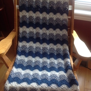 May include: A crocheted blanket with a wavy pattern in shades of blue and white. The blanket is draped over a wooden rocking chair.