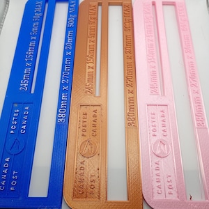 May include: Three plastic postal templates in blue, bronze, and pink. Each template has the text "CANADA POST" and measurements in millimeters and grams. The templates are rectangular with a cutout section.