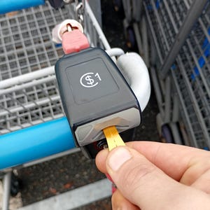 May include: A close-up of a shopping cart coin lock mechanism. The black mechanism has a white dollar sign and the number 1. A yellow token is being inserted into the slot to release the cart.