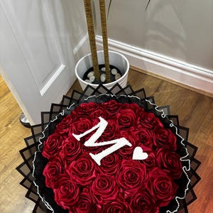 May include: A circular bouquet of red roses, featuring a white letter "M" and a heart. The arrangement is set in black and white decorative paper, creating a striking contrast.
