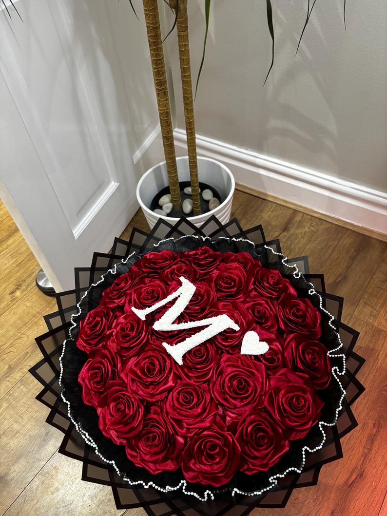 May include: A circular bouquet of red roses, featuring a white letter "M" and a heart. The arrangement is set in black and white decorative paper, creating a striking contrast.