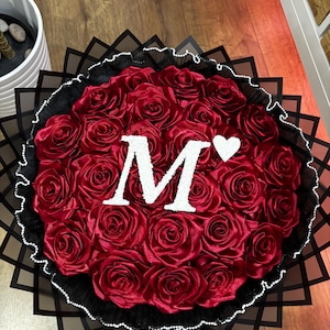May include: A circular bouquet of red satin roses, featuring a large white letter "M" and a white heart. The arrangement is set against a backdrop of black paper and a white beaded border.