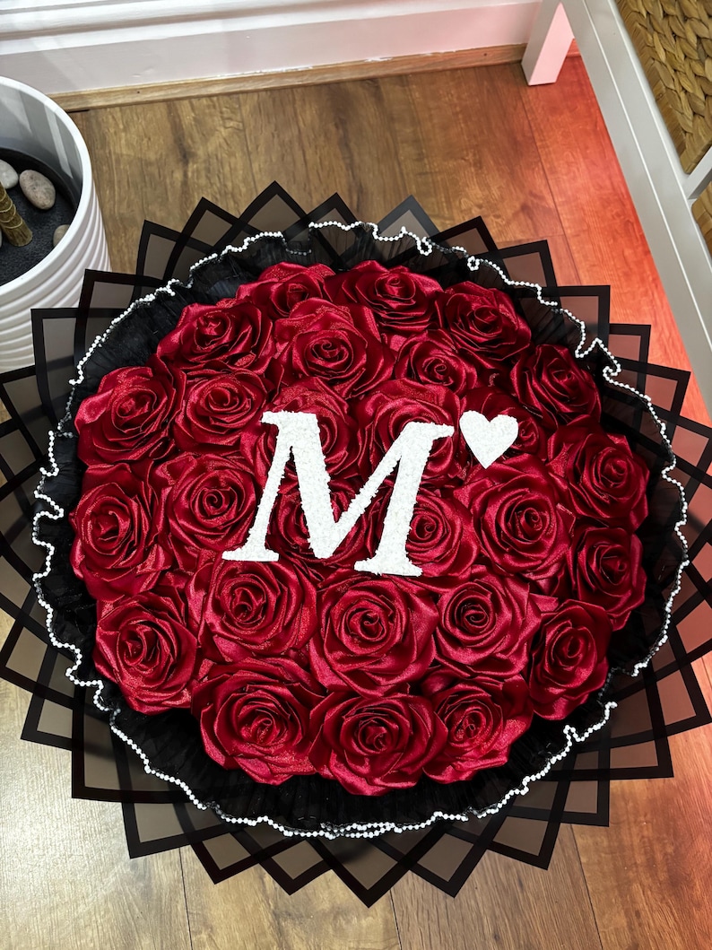 May include: A circular bouquet of red satin roses, featuring a large white letter "M" and a white heart. The arrangement is set against a backdrop of black paper and a white beaded border.