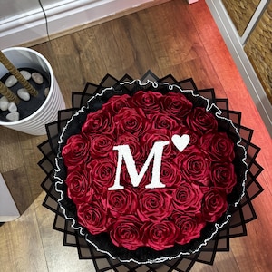 May include: A circular arrangement of red roses, featuring a white letter "M" and a heart. The bouquet is framed by a black decorative border and a white beaded trim, creating a striking visual.