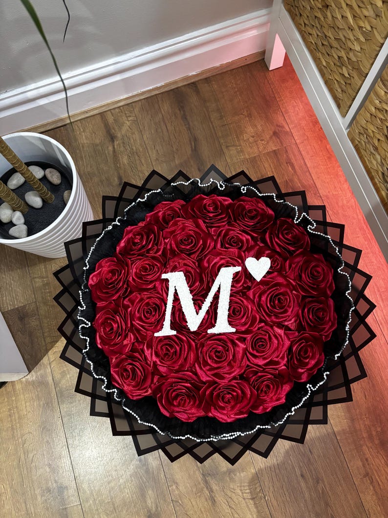 May include: A circular arrangement of red roses, featuring a white letter "M" and a heart. The bouquet is framed by a black decorative border and a white beaded trim, creating a striking visual.