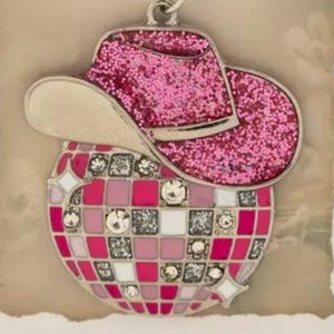 May include: A pink glitter cowboy hat charm sits above a disco ball design. The disco ball has a mosaic pattern of pink, white, and silver squares, with decorative gems. A silver-tone bail is attached for hanging.