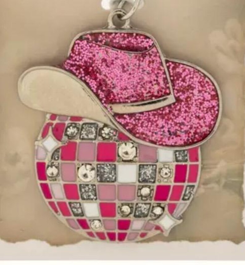 May include: A pink glitter cowboy hat charm sits above a disco ball design. The disco ball has a mosaic pattern of pink, white, and silver squares, with decorative gems. A silver-tone bail is attached for hanging.