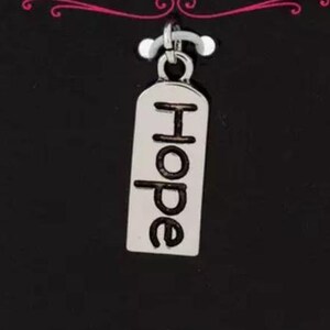 May include: A silver charm with the word "Hope" in black lettering. The charm is rectangular with rounded edges and hangs from a small silver loop. The background is black.