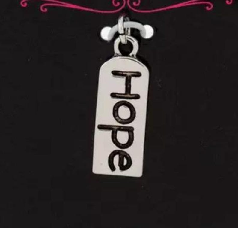 May include: A silver charm with the word "Hope" in black lettering. The charm is rectangular with rounded edges and hangs from a small silver loop. The background is black.