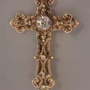 May include: An ornate gold-coloured cross pendant with a large, clear central gemstone and smaller accent stones. The cross features intricate scrollwork and a loop for hanging. A religious symbol.