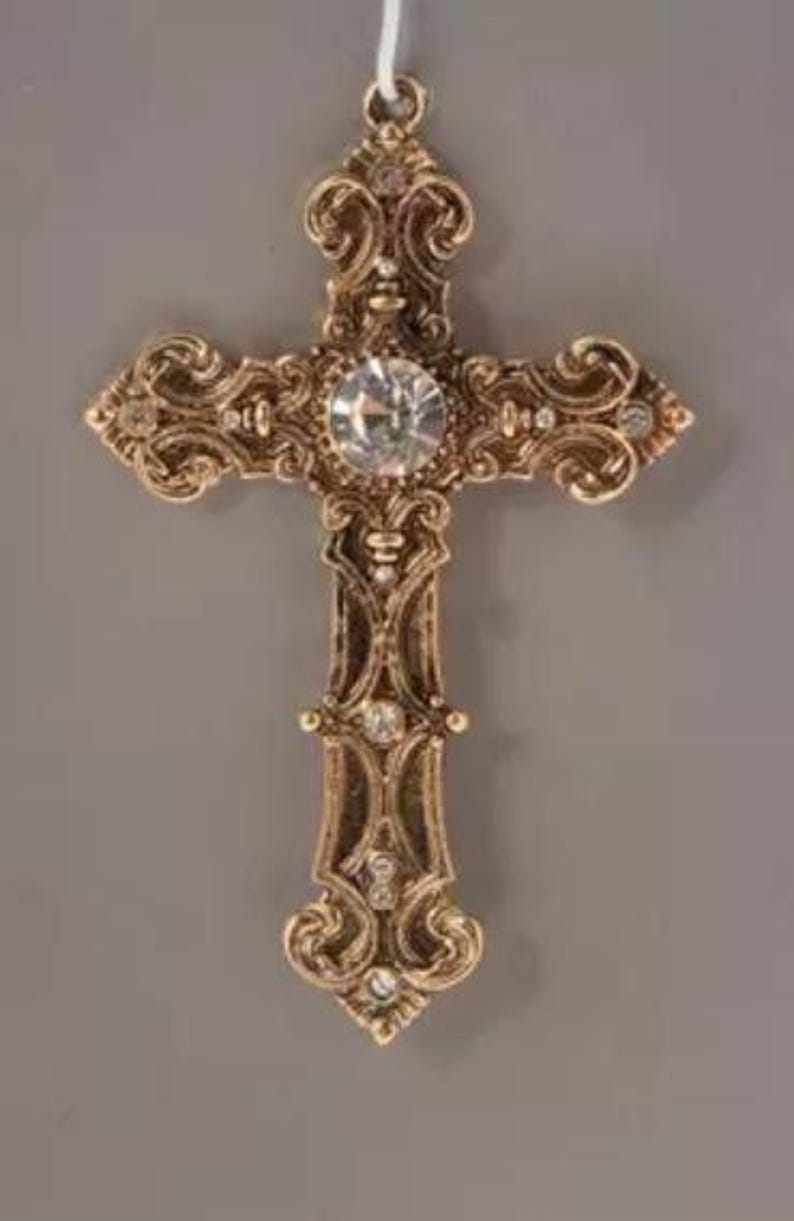 May include: An ornate gold-coloured cross pendant with a large, clear central gemstone and smaller accent stones. The cross features intricate scrollwork and a loop for hanging. A religious symbol.