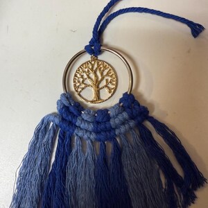 Puede incluir: Un colgante de pared de macramé con un colgante dorado de árbol de la vida dentro de un anillo plateado. El diseño utiliza tonos de azul, con una parte superior anudada y una parte inferior con flecos. Decoración de pared.