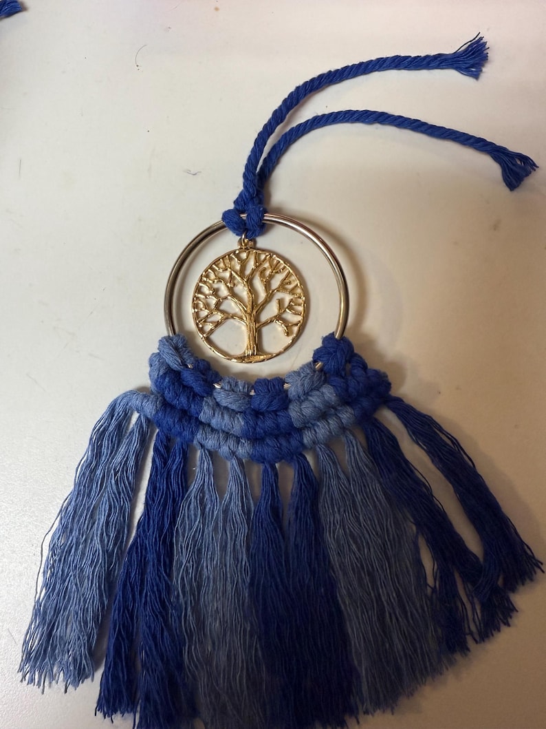 May include: A macrame wall hanging with a gold tree of life pendant within a silver ring. The design uses various shades of blue, with a knotted top and fringed bottom. The decorative hanging is designed to be displayed on a wall.