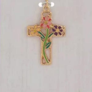 May include: Gold-coloured cross pendant with colourful floral accents. The cross features a stem with green leaves and flowers in pink, yellow, and purple. The pendant hangs from a delicate chain, set against a light background.