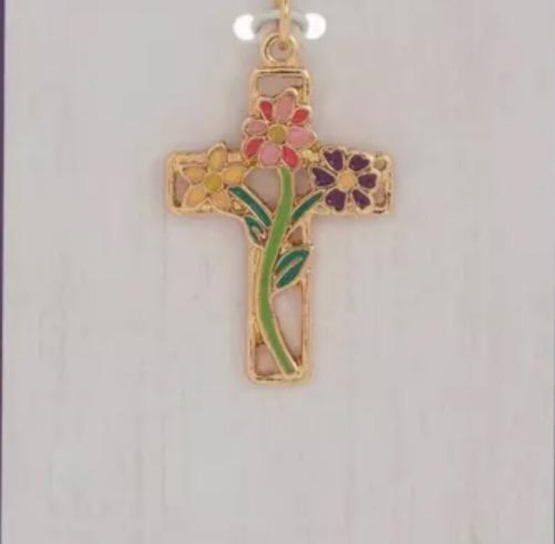 May include: Gold-coloured cross pendant with colourful floral accents. The cross features a stem with green leaves and flowers in pink, yellow, and purple. The pendant hangs from a delicate chain, set against a light background.