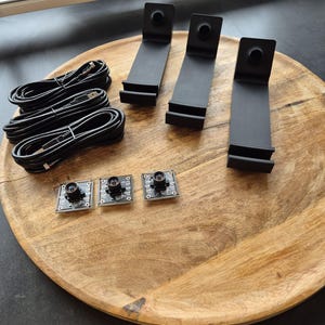 May include: Three black L-shaped plastic stands, three black USB cables coiled into circles, and three small square circuit boards with central camera lenses. The components are arranged on a round wooden surface with a visible grain pattern.