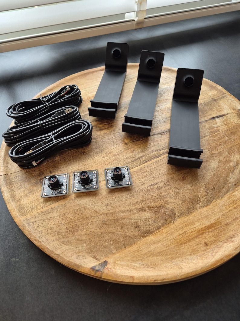 May include: Three black L-shaped plastic stands, three black USB cables coiled into circles, and three small square circuit boards with central camera lenses. The components are arranged on a round wooden surface with a visible grain pattern.