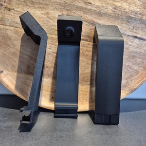 May include: Three black plastic camera mounts with a textured finish. The centre mount features a circular lens positioned at the top. Each mount has an angled top section and a flat, vertical body with a specialised clip mechanism at the base.