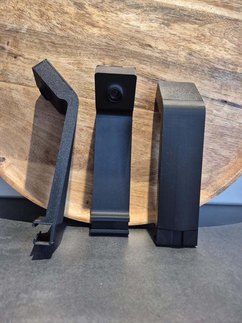 May include: Three black plastic camera mounts with a textured finish. The centre mount features a circular lens positioned at the top. Each mount has an angled top section and a flat, vertical body with a specialised clip mechanism at the base.