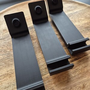 May include: Three matte black L-shaped stands, each featuring a circular camera lens mounted on the upper vertical section and a horizontal base with a grooved ledge designed to hold a device securely in an upright position.