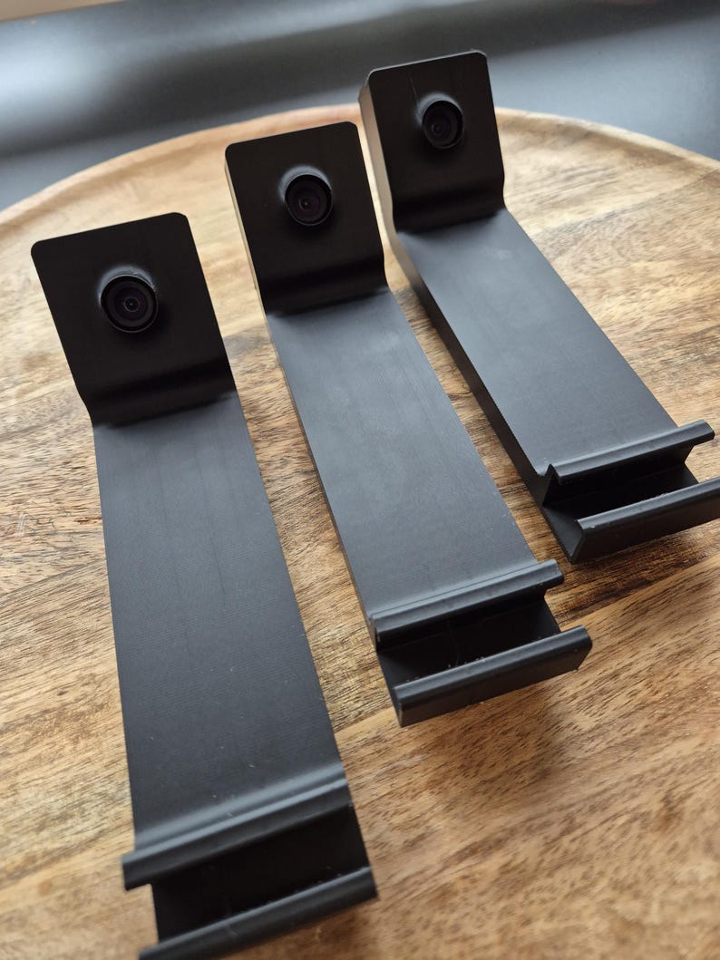 May include: Three matte black L-shaped stands, each featuring a circular camera lens mounted on the upper vertical section and a horizontal base with a grooved ledge designed to hold a device securely in an upright position.