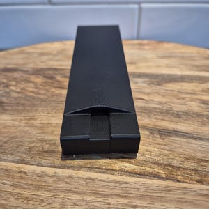May include: A matte black rectangular object with a smooth finish and a central, recessed notch at the front edge. The item features a long, narrow, and flat top surface that tapers slightly towards the front, constructed from a solid material.
