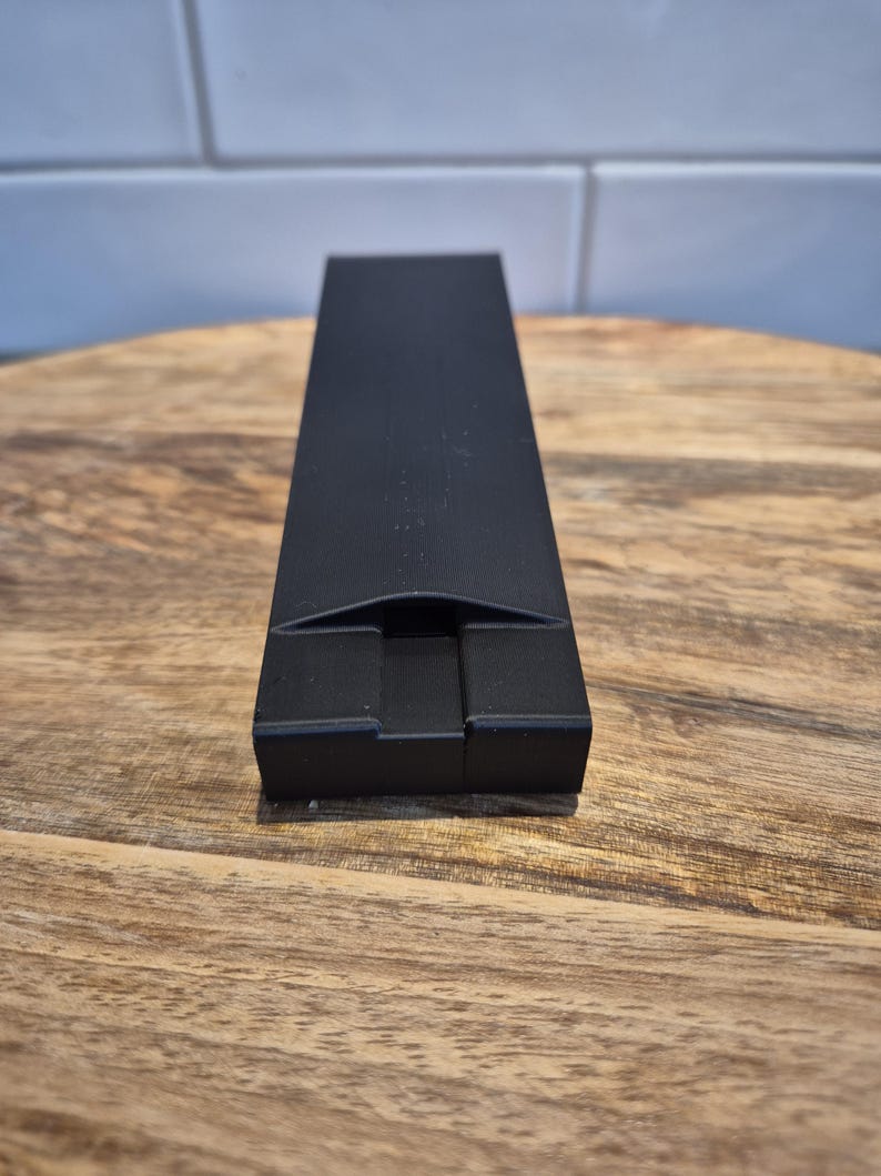 May include: A matte black rectangular object with a smooth finish and a central, recessed notch at the front edge. The item features a long, narrow, and flat top surface that tapers slightly towards the front, constructed from a solid material.