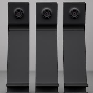 May include: Three matte black vertical stands, each featuring a circular camera lens mounted at the top and a horizontal ledge at the base. The devices have a uniform, elongated rectangular shape with a slight bend near the top section.