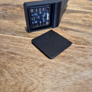 May include: A black 3D-printed plastic housing with an L-shaped arm and an exposed square circuit board featuring various electronic components and a white connector port, accompanied by a separate matching black square cover plate.