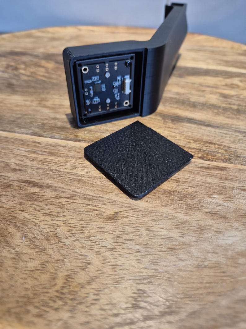 May include: A black 3D-printed plastic housing with an L-shaped arm and an exposed square circuit board featuring various electronic components and a white connector port, accompanied by a separate matching black square cover plate.