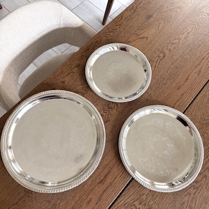 May include: Three silver-colored serving trays of varying sizes are arranged on a wooden table. Each tray has a decorative, beaded edge. The trays are round and reflect the light, creating a bright and elegant appearance. The table has a rich, dark wood grain.