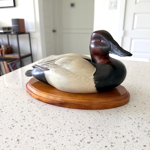 May include: A hand-carved wooden duck decoy featuring a dark brown head with a small red eye, a black chest, and a light cream-colored body. The duck is mounted on a smooth, oval-shaped wooden base with a polished finish and visible wood grain.