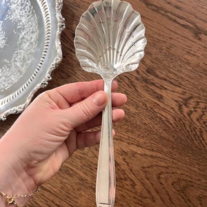 May include: A silver serving spoon with a shell-shaped bowl and a long, tapered handle. The spoon is held against a wooden surface, with a decorative silver tray in the background. The spoon is likely part of a vintage or antique silverware set.