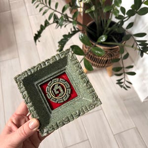 May include: A square, green trinket dish with a raised, textured border. The center features a red square with a gold-colored circular design. The dish is held in a hand, with a plant and wooden shelf in the background.
