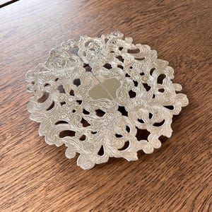 May include: A silver-colored, ornate coaster with a detailed, openwork design. The coaster features a symmetrical, floral pattern with a central, shield-shaped area. The coaster is circular and rests on a wooden surface.
