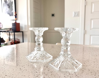 May include: Two clear crystal candle holders with a square base and a detailed, faceted design. The candle holders have a classic, elegant shape, perfect for adding a touch of sophistication to any space. They are displayed on a light-colored countertop.