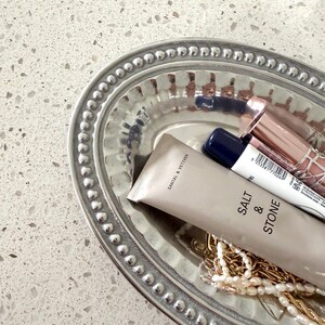 Puede incluir: Una bandeja ovalada plateada con borde de cuentas contiene productos de belleza y joyas. Incluye un tubo de loci&oacute;n "SALT & STONE", barras de labios y un collar de oro. La bandeja est&aacute; sobre una superficie blanca moteada.