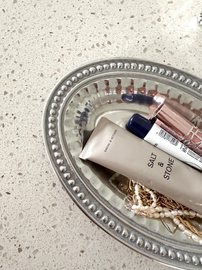 Puede incluir: Una bandeja ovalada plateada con borde de cuentas contiene productos de belleza y joyas. Incluye un tubo de loci&oacute;n "SALT & STONE", barras de labios y un collar de oro. La bandeja est&aacute; sobre una superficie blanca moteada.