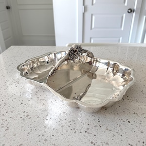 May include: A silver-colored metal serving dish with a rectangular shape, scalloped edges, and a central arched handle featuring ornate floral detailing. The surface of the dish is reflective and smooth, divided into two sections by the handle.
