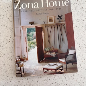 May include: A book titled "Zona Home" with the subtitle "Essential Designs for Living" by Louis Sagar. The cover features a photograph of an interior space with open doors leading to a garden, and various furniture pieces.