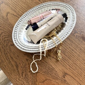 Puede incluir: Una bandeja ovalada plateada con borde de cuentas contiene productos de belleza y joyas. La bandeja contiene un tubo de crema, un l&aacute;piz labial, un tubo negro, una cadena dorada y un collar de perlas. Los art&iacute;culos est&aacute;n dispuestos sobre una superficie de madera.