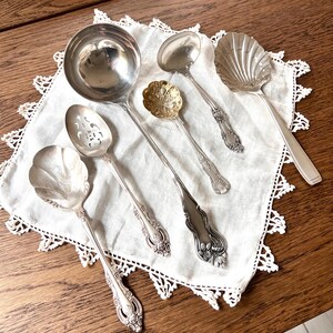 May include: An assortment of antique silver serving spoons and ladles, including a large ladle, a slotted spoon, and a shell-shaped spoon, arranged on a white lace-edged cloth. The spoons have ornate handles and are displayed on a wooden surface.